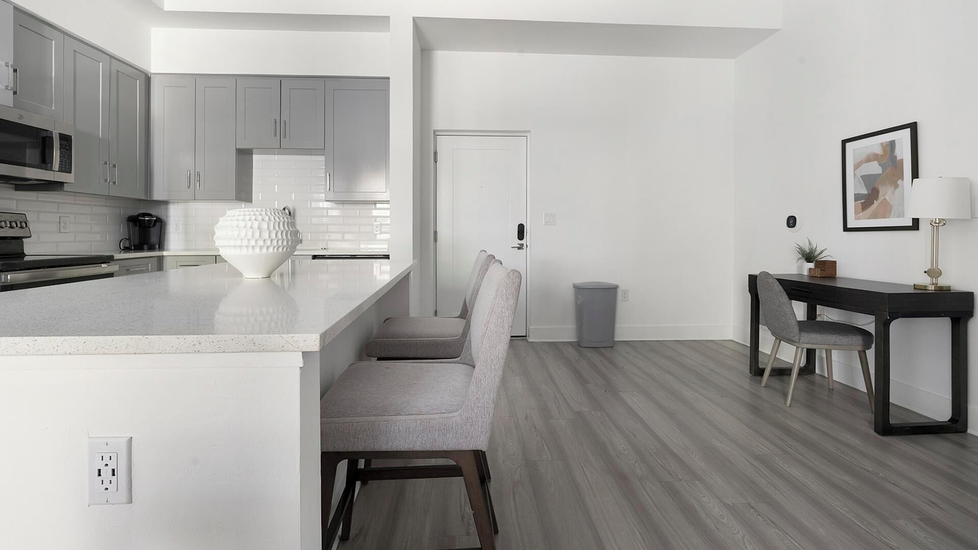 Una cocina moderna con armarios grises, una encimera blanca y un microondas empotrado. Se observa una pequeña isla con un bol blanco y una silla. Al fondo, un comedor con una mesa y sillas.
