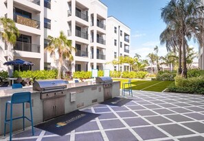 A patio area with a grill station, outdoor seating, and a view of a multi-story building with balconies. - Landing | Stunning 1BD, Gym, Game Room (Fort Myers)