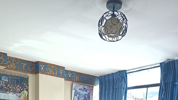 A hotel room with a large bed, a ceiling light fixture, a window with blue curtains, and two wall paintings. - Samana Wasi Urpicha (Cusco)