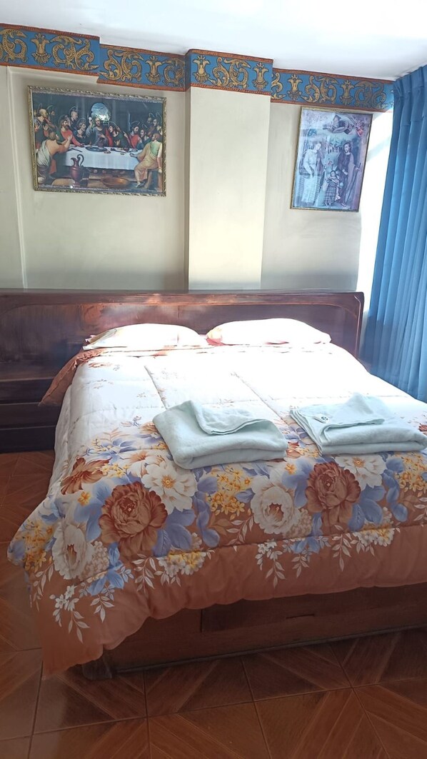 Une chambre d’hôtel avec un grand lit, une couvre-lit à motifs floraux, deux serviettes, une tête de lit en bois et deux œuvres d’art encadrées accrochées au mur.