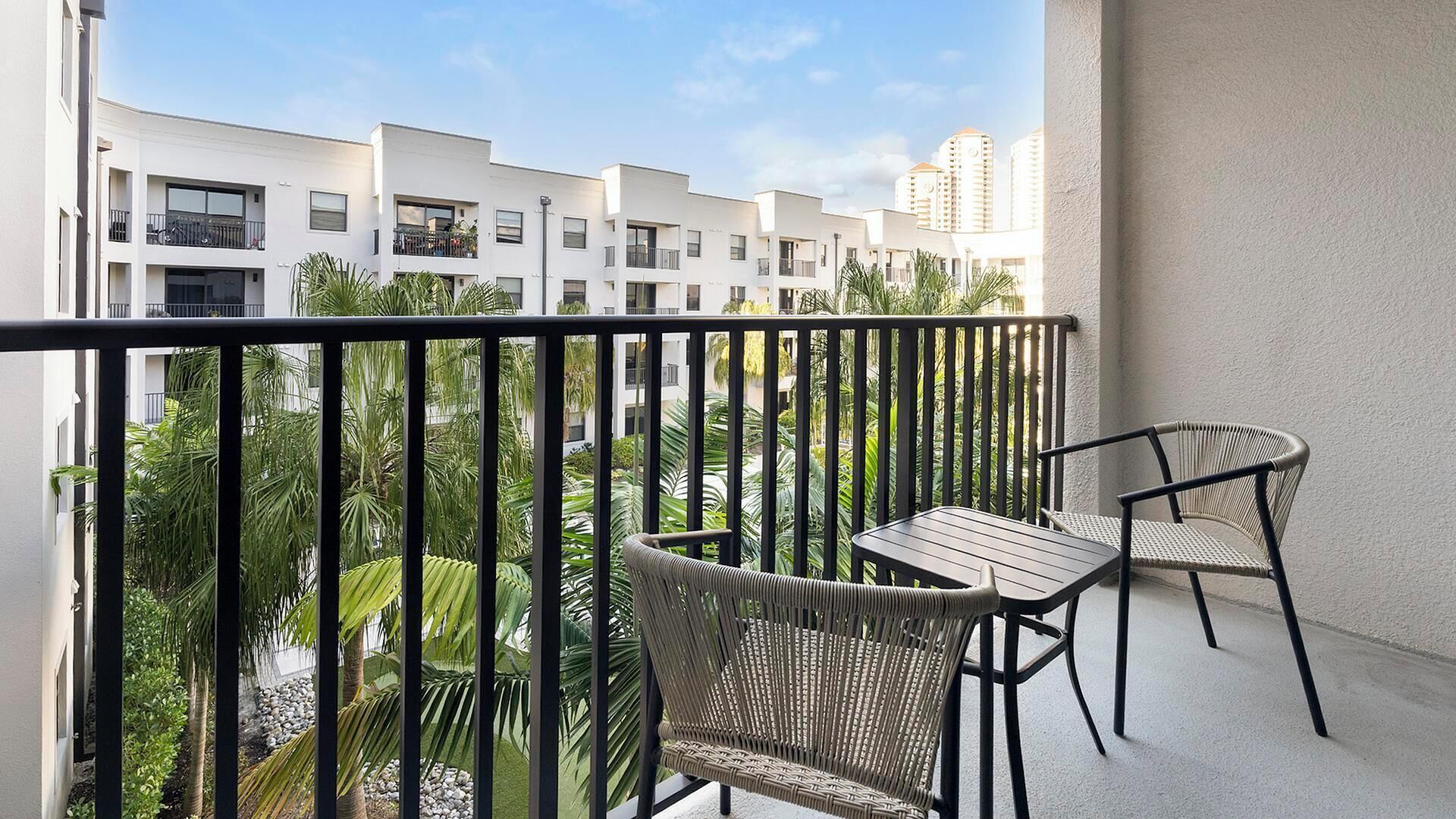 Un balcon avec deux fauteuils en osier et une petite table, donnant sur un quartier résidentiel avec des bâtiments blancs et des palmiers.