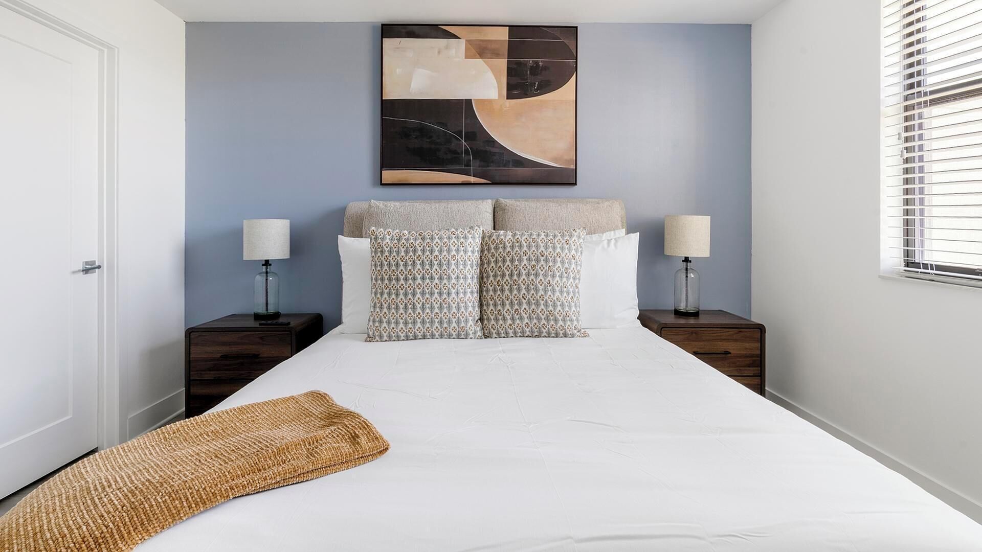 A bedroom with a bed, two nightstands, a lamp on each, a patterned pillow, a plain pillow, and a woven throw.
