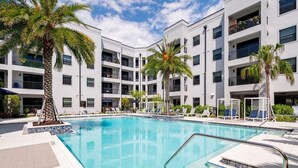 Ein modernes Hotel mit einem zentralen Swimmingpool, mehreren Balkonen und Palmen.