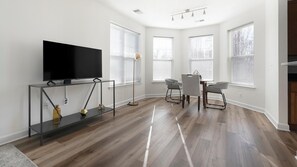 Ein modernes Wohnzimmer mit einem Flachbildfernseher auf einem schwarzen Konsolentisch, einem Essbereich mit kleinem Tisch und Stühlen sowie großen Fenstern mit Jalousien.