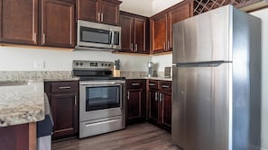 Una cocina con armarios de madera oscura, electrodomésticos de acero inoxidable, incluyendo un refrigerador, horno y microondas, y una encimera de granito.