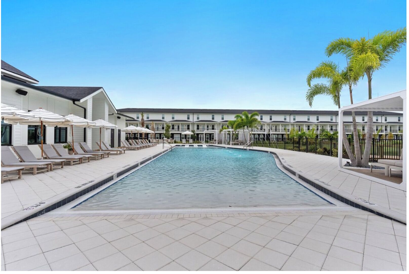 A swimming pool with lounge chairs and umbrellas, surrounded by a building with multiple windows and a tiled deck.