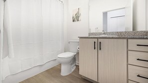 A bathroom with a toilet, a shower curtain, a sink with a granite countertop, and wooden cabinets. - Landing | Amazing 2BD, Clubhouse, Pool (Lakeland)