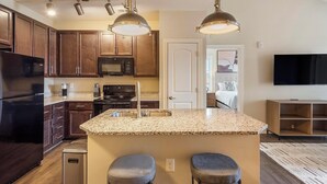 A modern kitchen with dark wood cabinets, a black refrigerator, a microwave, and a stove. There is a granite countertop with a sink and two bar stools. A television is mounted on the wall in the adjacent room. - Landing | Modern 2BD, Clubhouse, Gym (Greer)