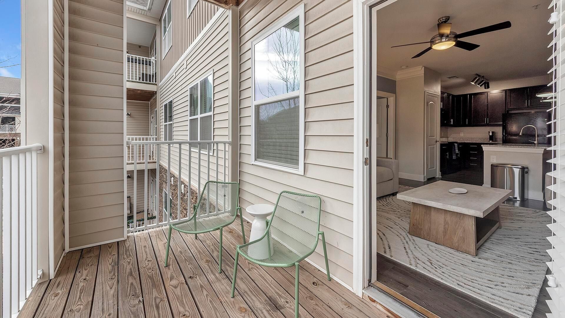 A wooden deck with two green chairs and a small table, a balcony with railings, and a kitchen area with dark cabinets and a countertop.