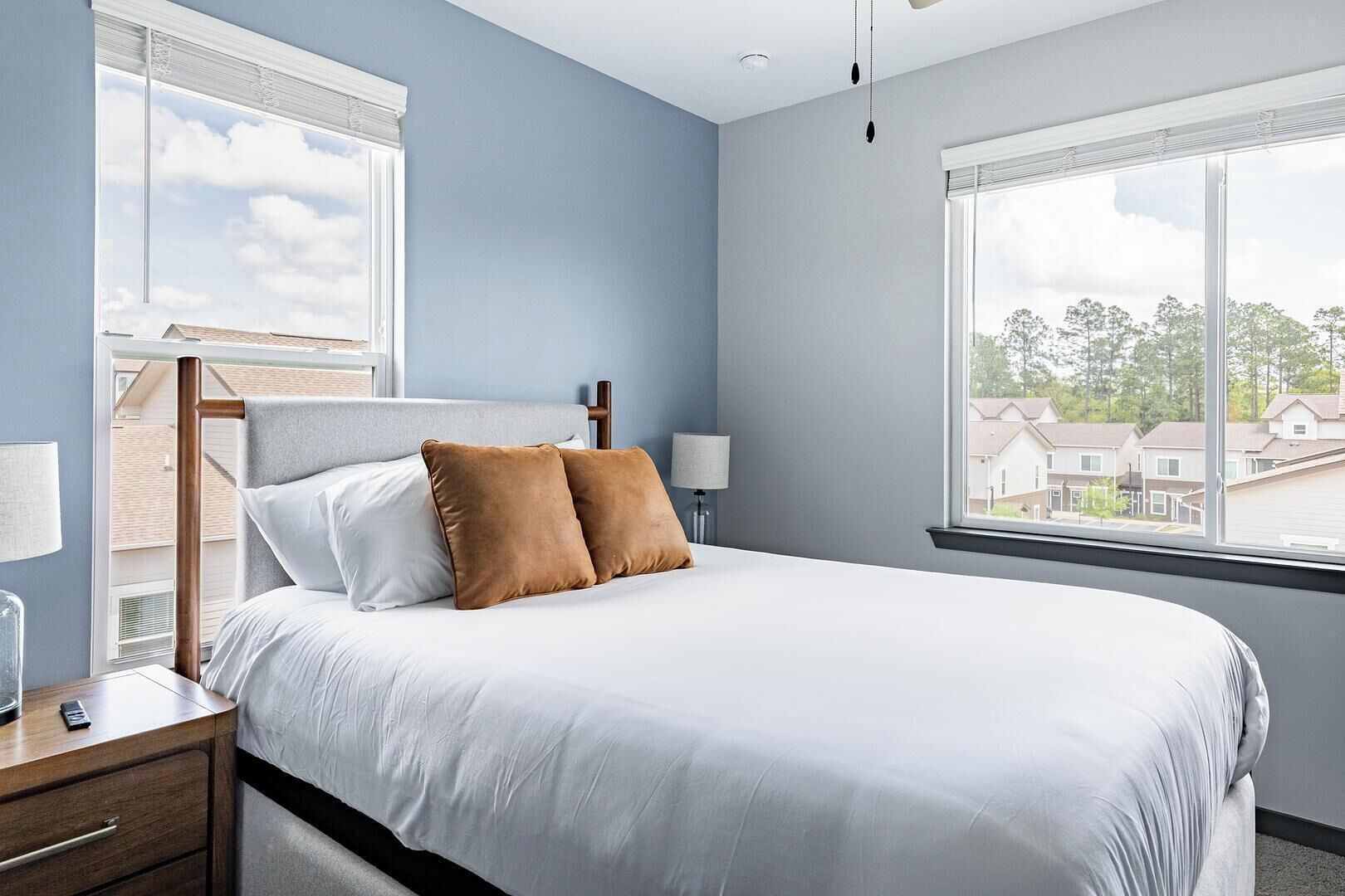 A bedroom with a bed, a nightstand, a lamp, and a window with a view of houses.