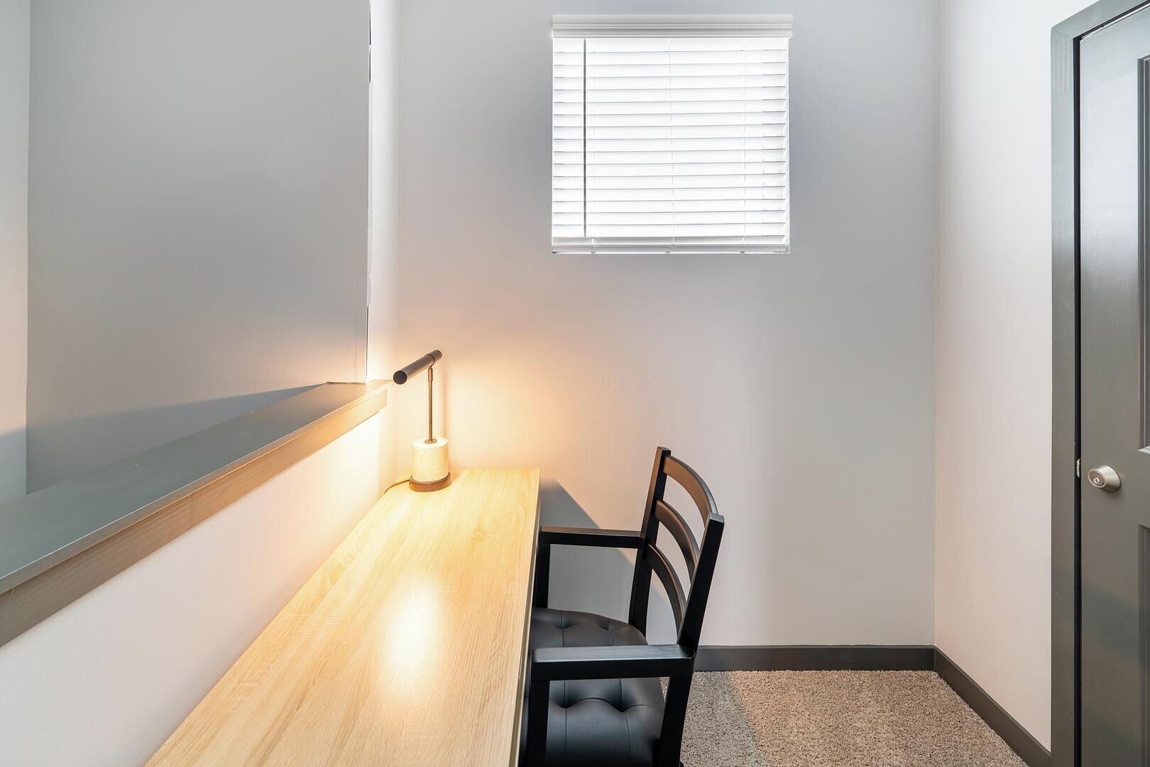 A small, well-lit room with a wooden table, a single black chair, a window with blinds, and a door.
