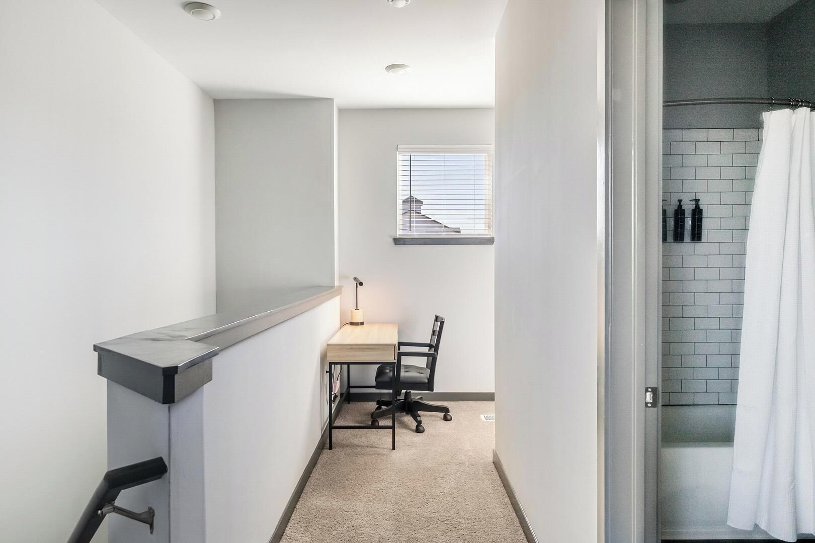 A narrow hallway with a desk and chair, a window with blinds, and a shower area with a white curtain.