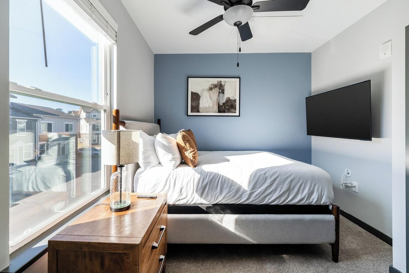 Un dormitorio con cama, mesita de noche, lámpara, ventilador de techo, televisor montado y ventana con vistas a edificios.