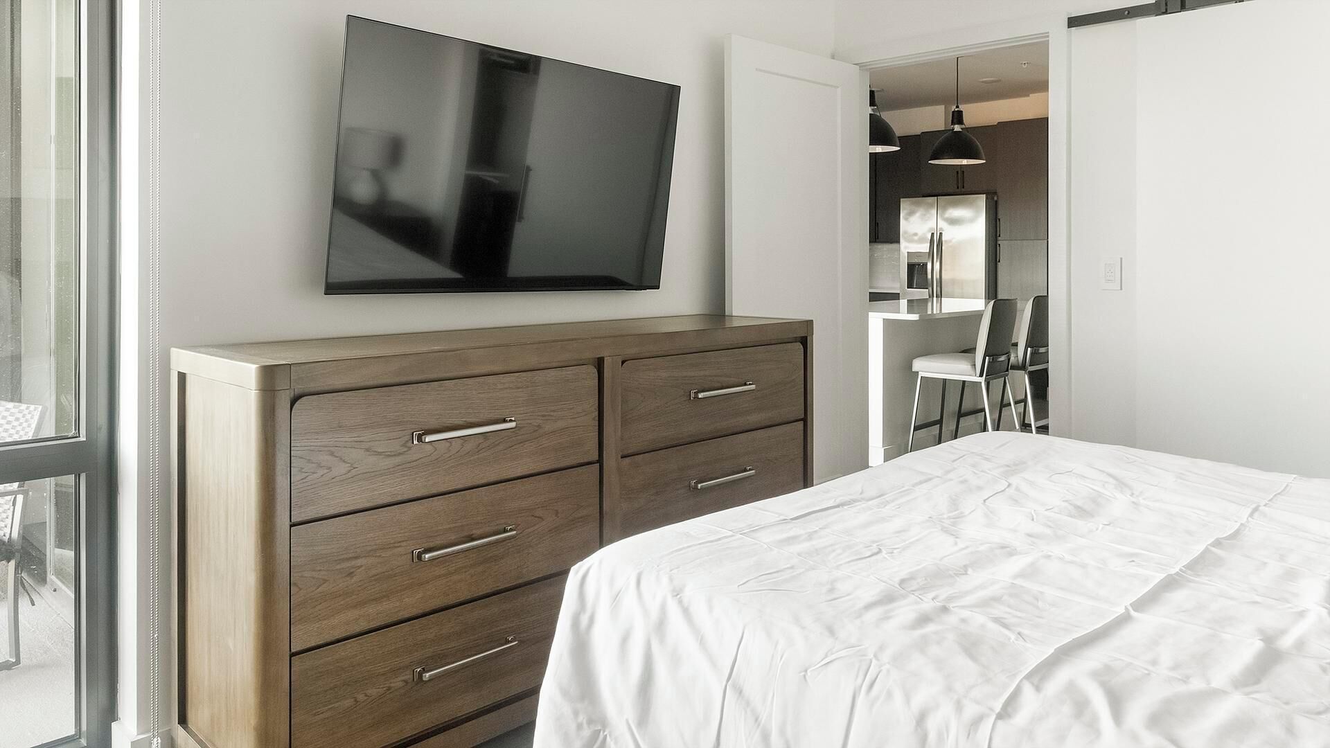 Un dormitorio con una cama grande, un aparador de madera con cajones, un televisor de pantalla plana montado en la pared, y vista al área de comedor contigua.