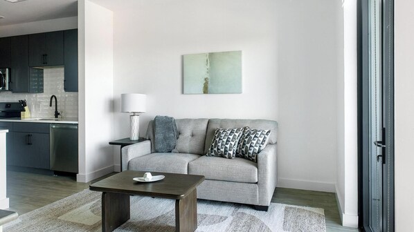 A modern living room with a grey sofa, a small table, and a lamp. There is a kitchen area with dark cabinets and a stainless steel refrigerator. - Landing | Amazing 1BD in Uptown Charlotte (Charlotte)