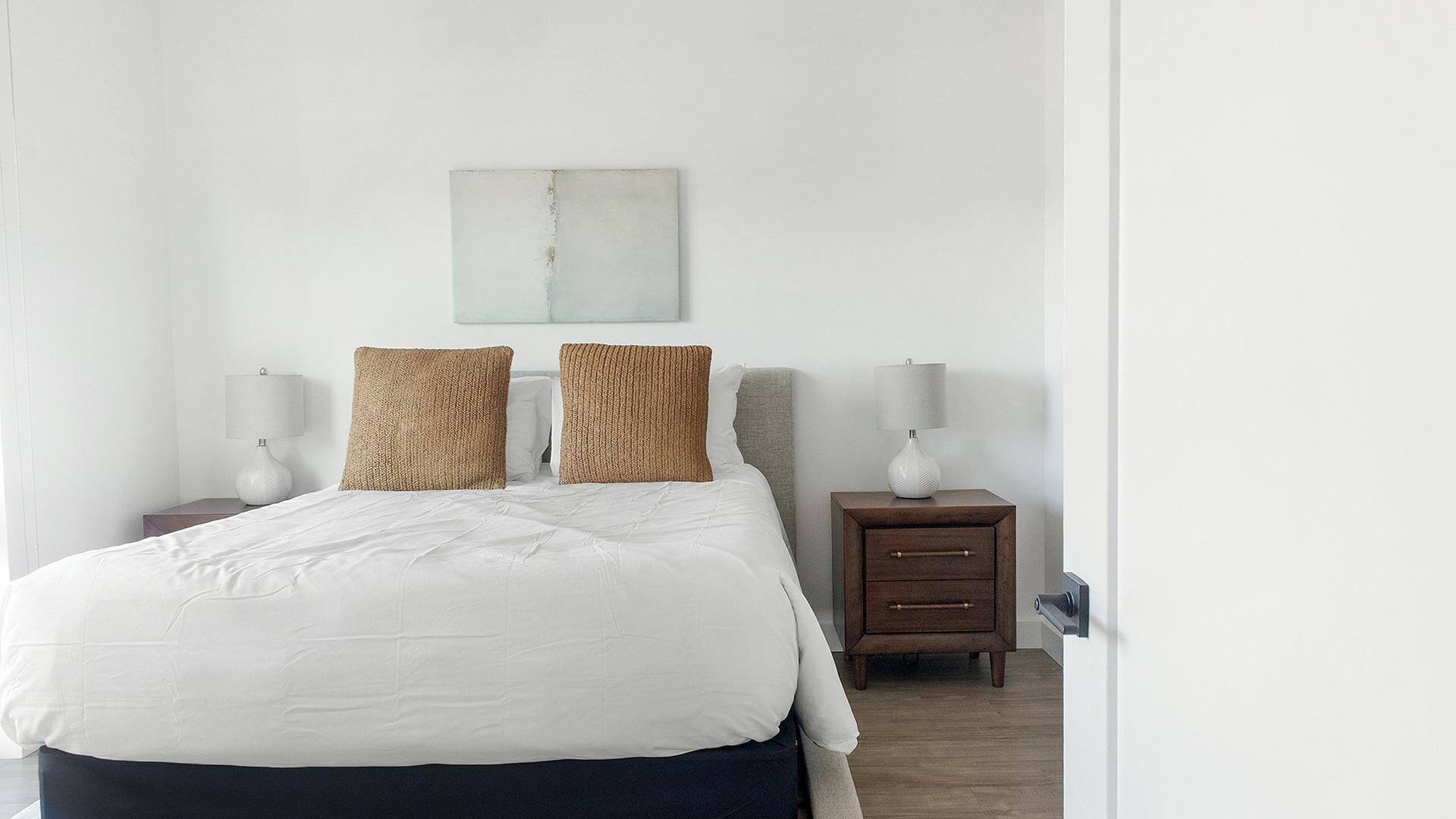 A neatly made bed with white bedding, two textured pillows, a bedside table with a lamp, and a small wooden nightstand with a drawer.