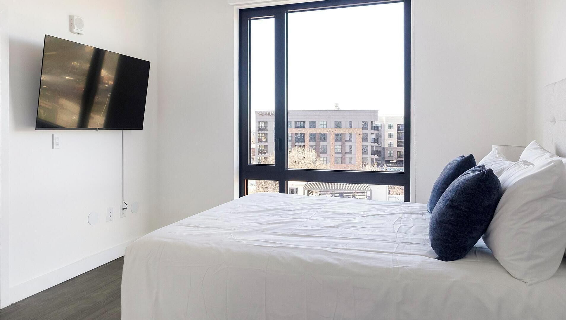 Un dormitorio con un ventanal grande, una cama con almohadas blancas y azul oscuro, y un televisor de pantalla plana montado en la pared.