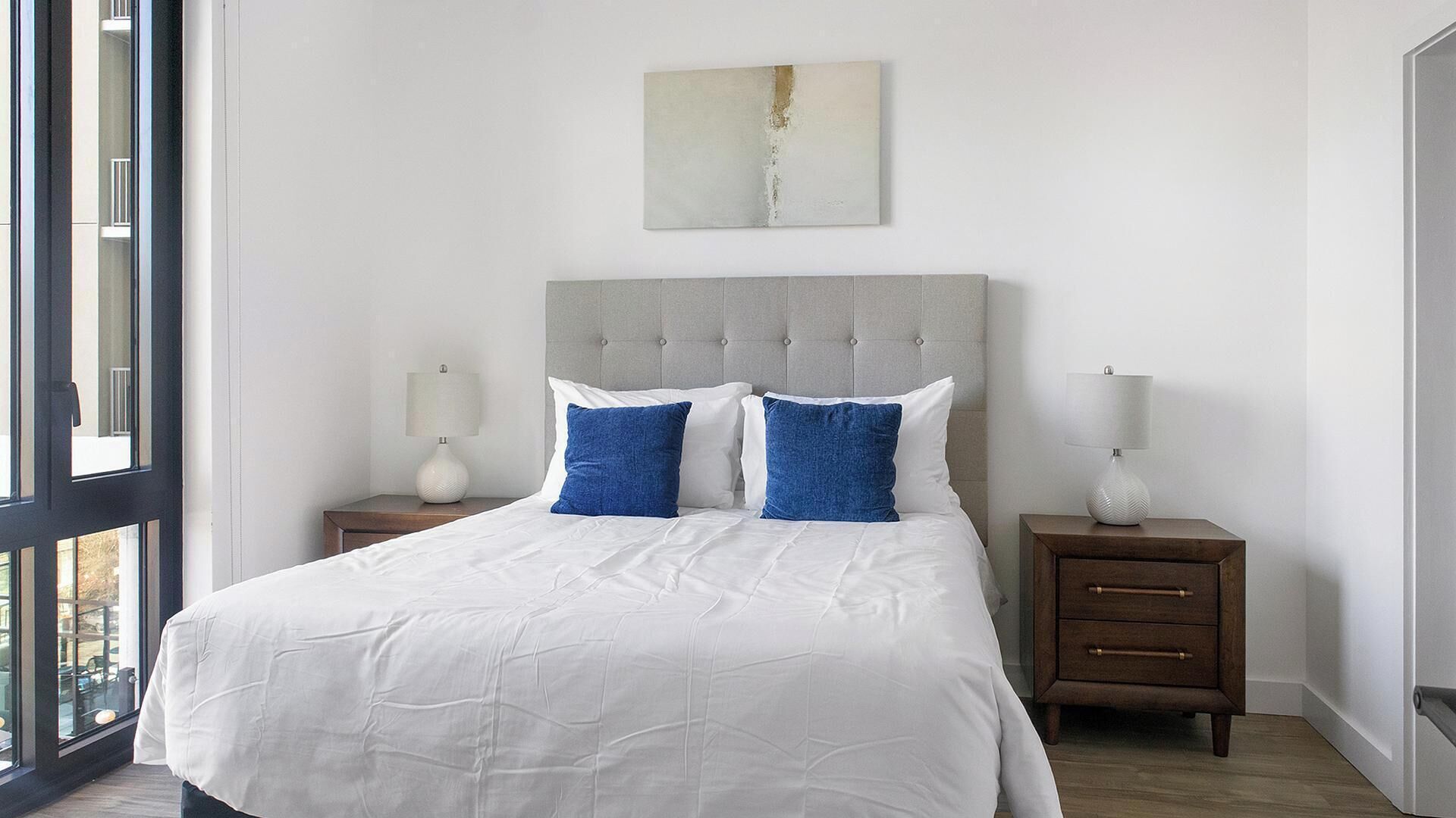 A bedroom with a large bed, two nightstands, a grey upholstered headboard, and a window with a view.