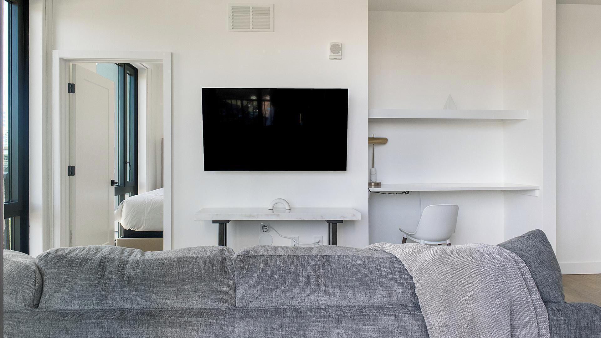 Un salon moderne avec un canapé gris, un téléviseur fixé au mur, une étagère blanche fixée au mur et une porte donnant sur une autre pièce.
