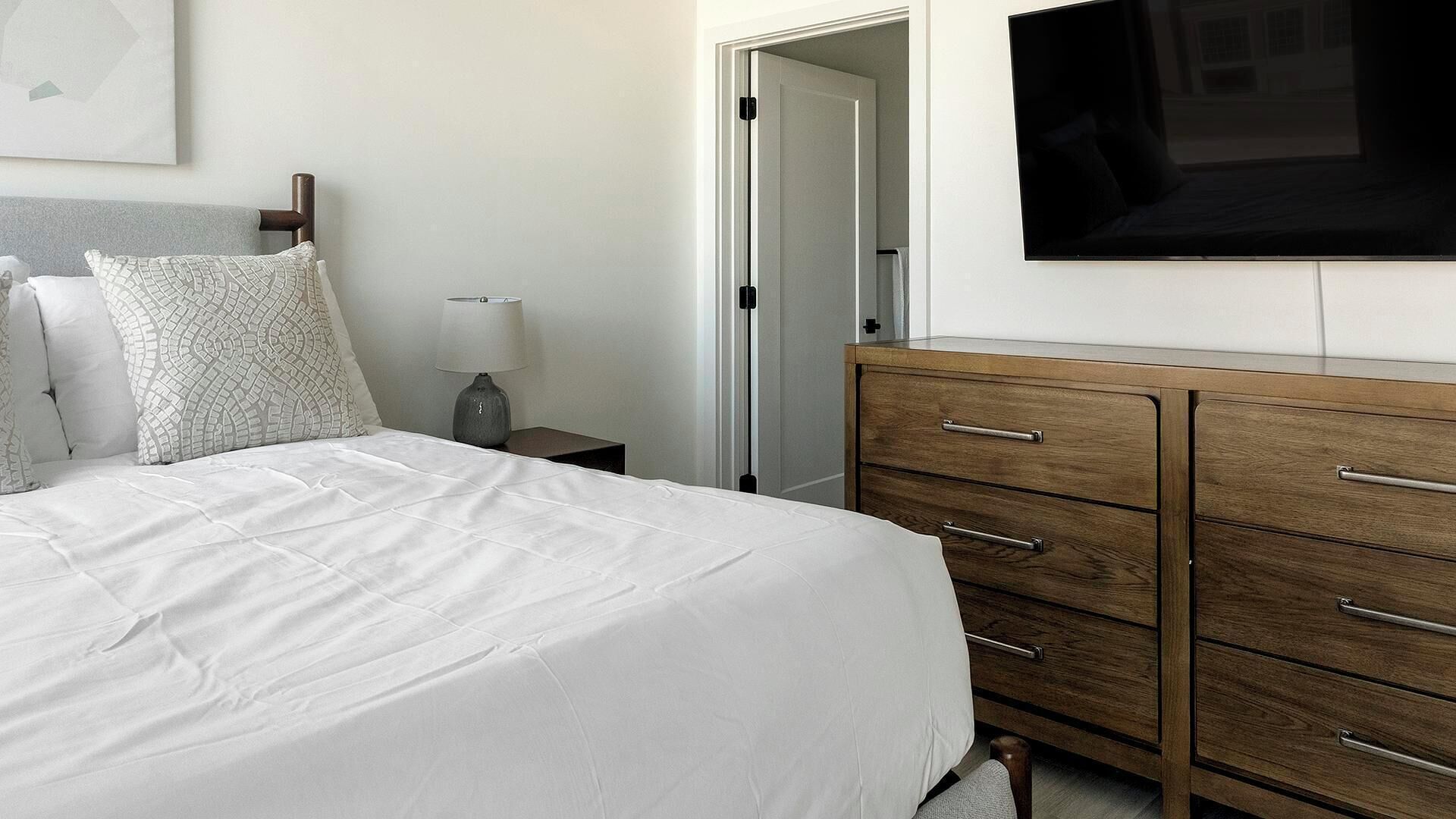 Un lit bien fait, avec du linge de lit blanc, une commode en bois dotée de tiroirs, une lampe de chevet et un téléviseur à écran plat fixé au mur.