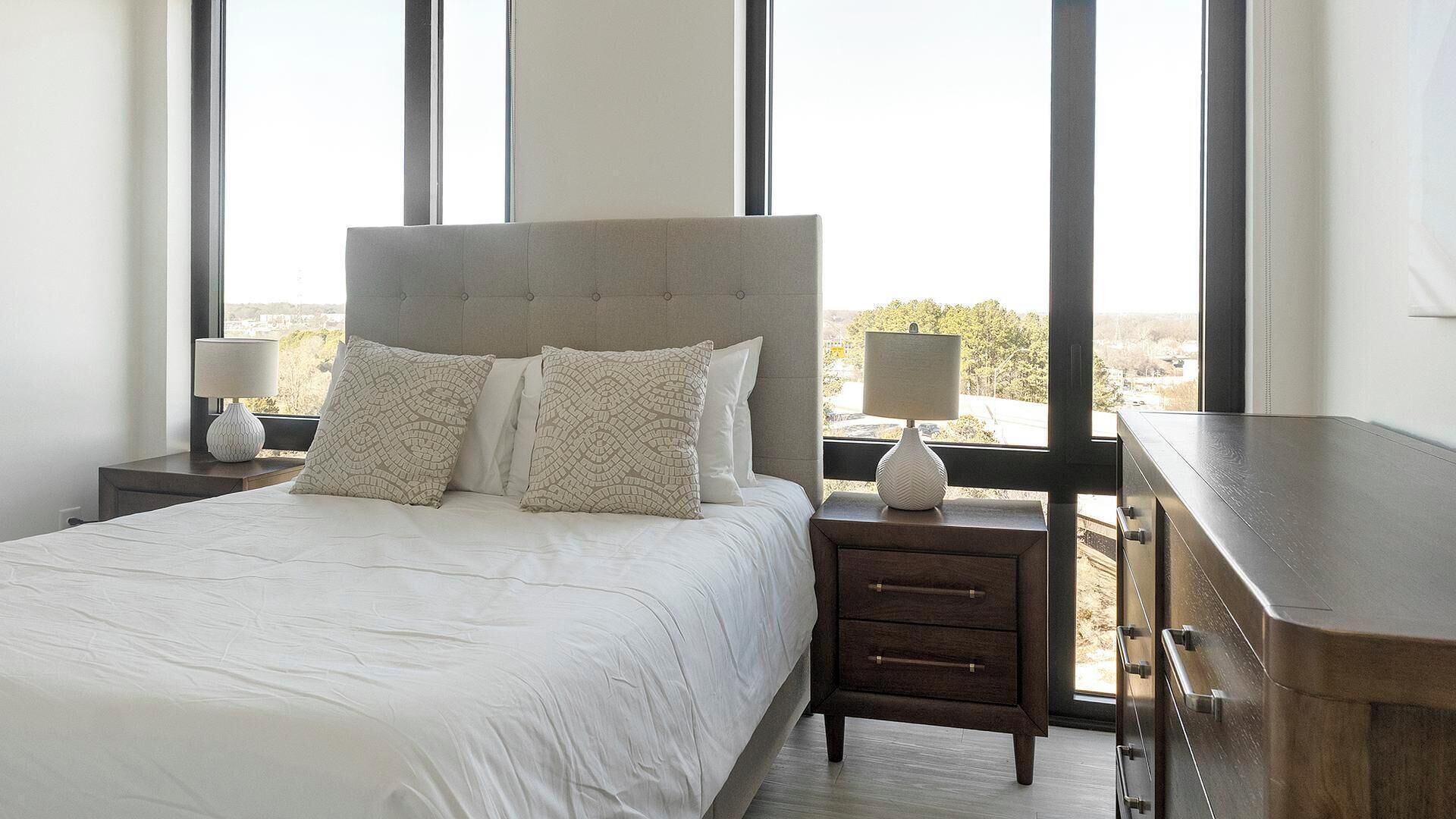 A bedroom with a large bed, two bedside tables with lamps, and a view of the outdoors through large windows.