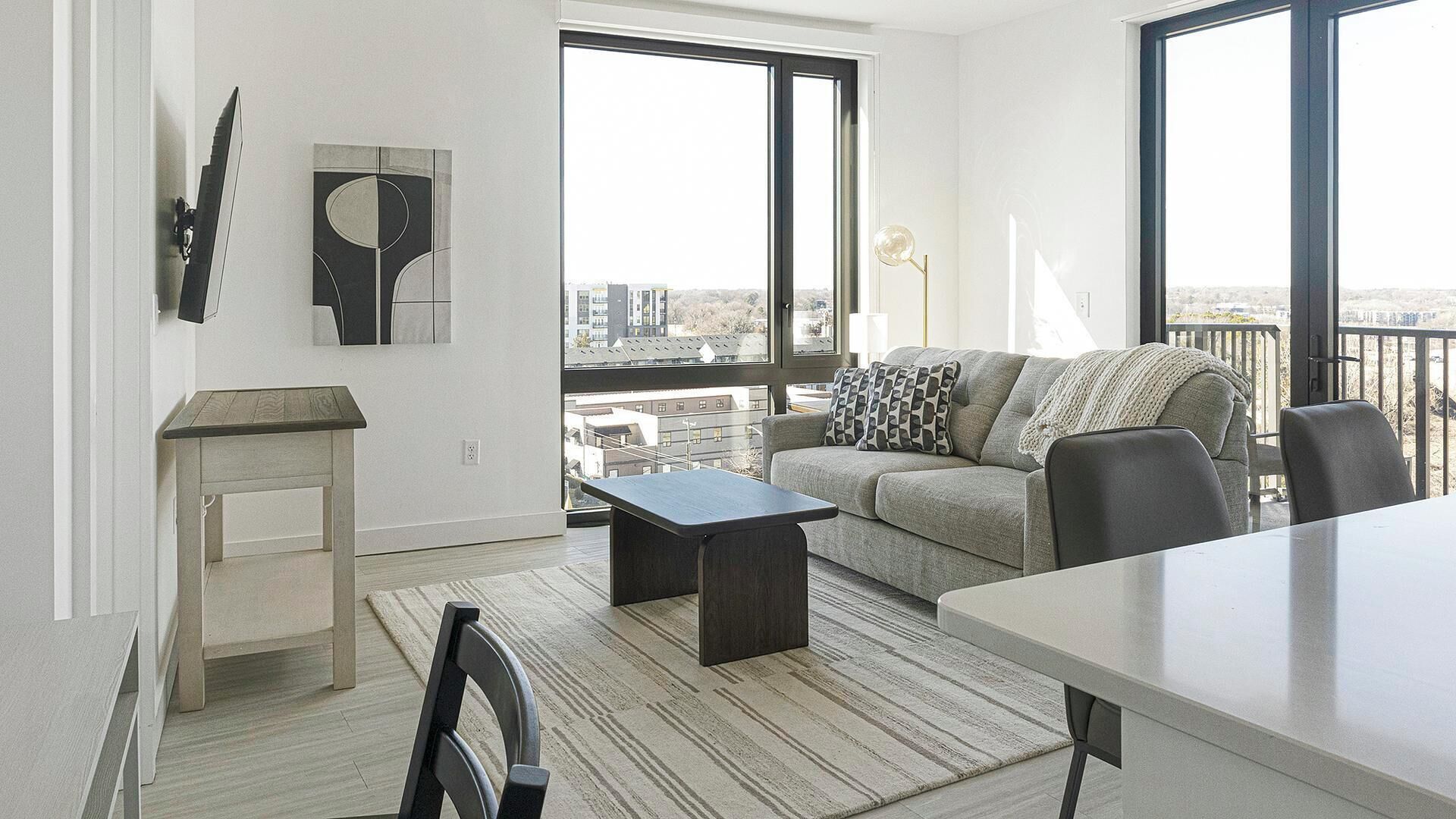 A modern living room with a sofa, a coffee table, and a dining table. There is a large window providing a view of the city.