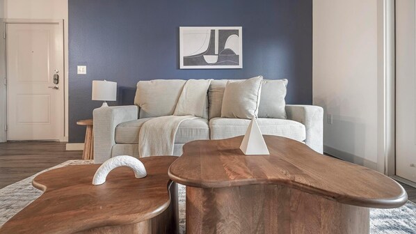 A modern living room with a dark blue accent wall, a white sofa with striped cushions, and a wooden coffee table with a white sculpture. - Landing | Modern 2BD, Gym, Game Room (Buda)
