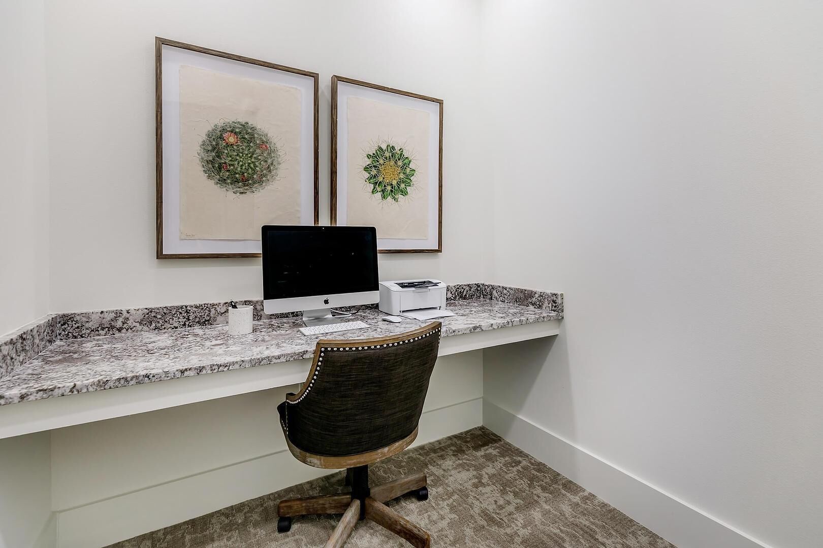 Una oficina en casa con un ordenador de escritorio, obras de arte enmarcadas, una encimera de mármol y una silla de asiento tejido oscuro.