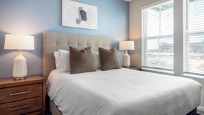 A neatly made bed with white bedding and two decorative pillows, flanked by nightstands with lamps, in a room with a large window and a piece of abstract art above the bed. - Landing | Incredible 2BD, Gym, Pool (Charlotte)