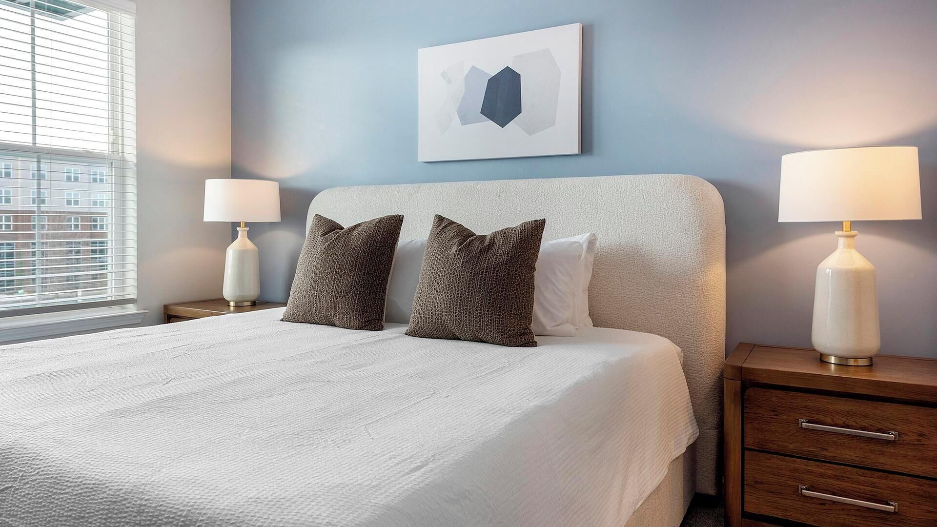 A neatly made bed with a white comforter and two decorative pillows, flanked by two bedside tables with lamps, in a room with a blue accent wall and a framed artwork above the bed.