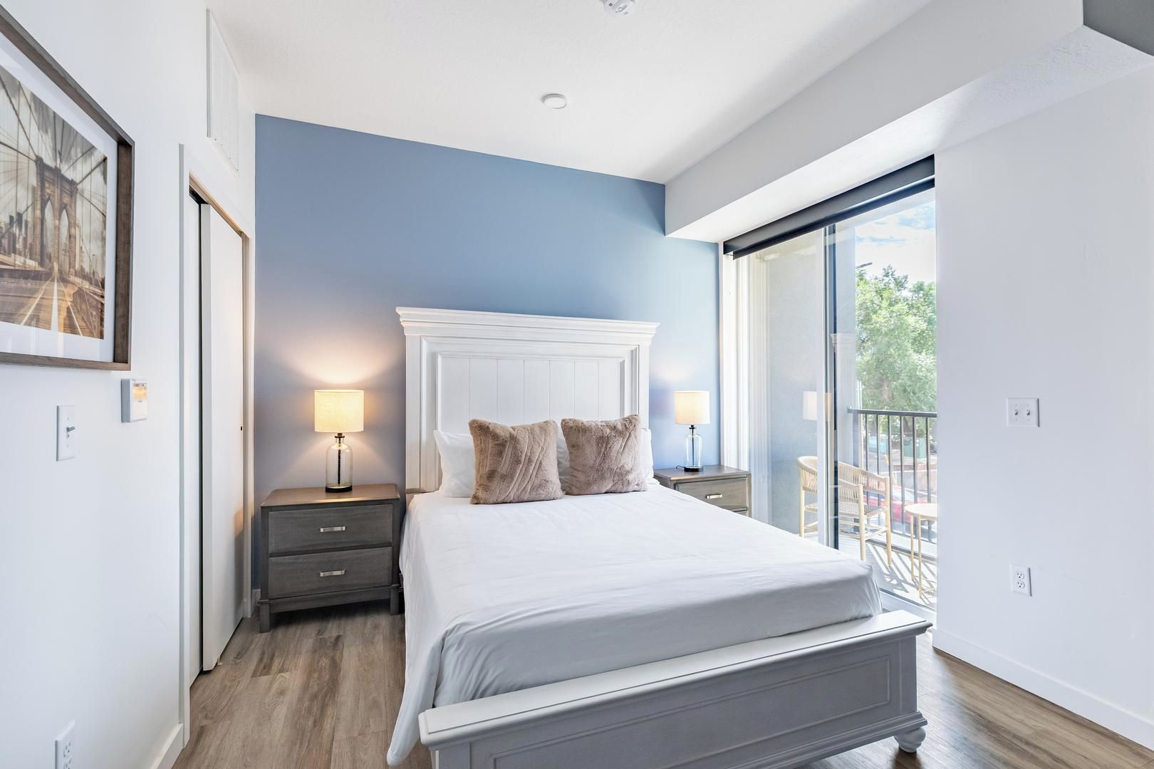 A bedroom with a large bed, two bedside tables with lamps, a nightstand, a framed picture on the wall, and a balcony with a view.