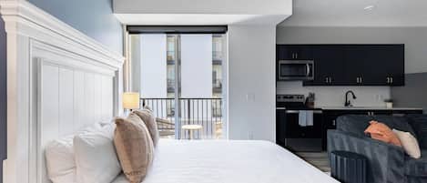 A modern hotel room with a large bed, a sofa, a kitchen area, and a balcony with a view.