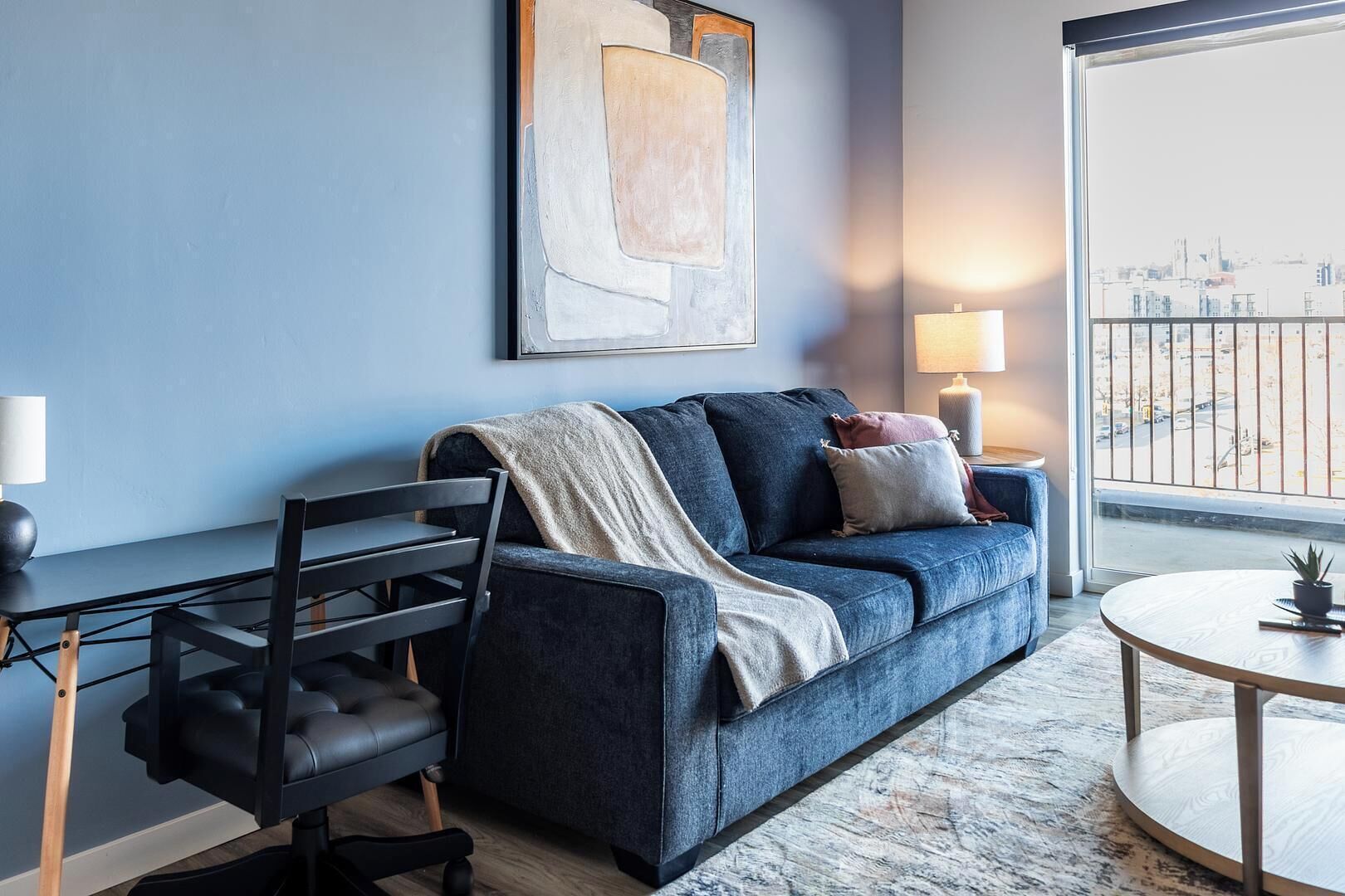 Una sala moderna con un sofá azul oscuro, un sillón negro, una mesa redonda de madera y un ventanal con vistas a la ciudad.