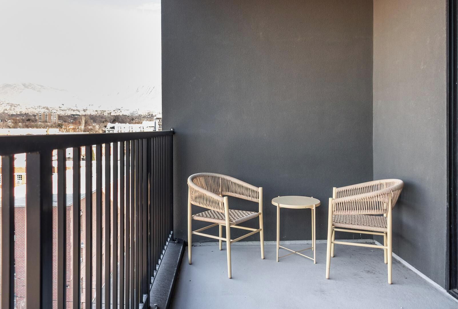 Un balcon avec deux fauteuils en osier et une petite table ronde, une rambarde en métal noir, et une vue sur un paysage urbain avec des bâtiments et des montagnes en arrière-plan.