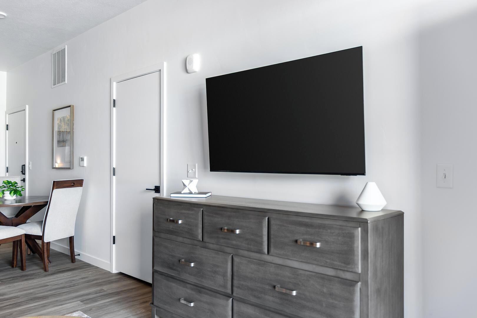 A modern room with a large flat-screen TV mounted on the wall, a grey dresser with drawers, a white lamp, and a dining area with a table and chairs.