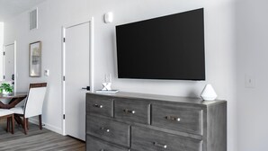 A modern room with a large flat-screen TV mounted on the wall, a grey dresser with drawers, a white lamp, and a dining area with a table and chairs. - Landing | Modern 1BD in Central City (Salt Lake City)
