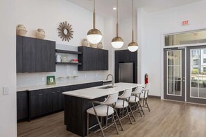 A modern kitchen with dark cabinetry, a central island with chairs, pendant lighting, and a view to the outside through a glass door.