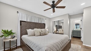A bedroom with a bed, a ceiling fan, a nightstand with a plant, and a bathroom visible through an open door.