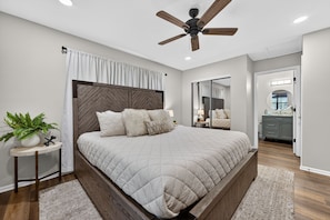 Quarto com cama, ventilador de teto, mesa de cabeceira com planta e banheiro visível através de uma porta aberta.