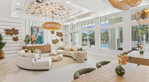 A spacious living room with a curved sofa, a coffee table, and a dining area with a wooden table and chairs. The room features large windows, a ceiling with a geometric pattern, and decorative wall art. - Landing | Chic 2BD, Clubhouse, Pool (Palm Bay)