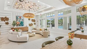 A spacious living room with a curved sofa, a coffee table, and a dining area with a wooden table and chairs. The room features large windows, a ceiling with a geometric pattern, and decorative wall art. - Landing | Chic 2BD, Pool, Clubhouse (Palm Bay)