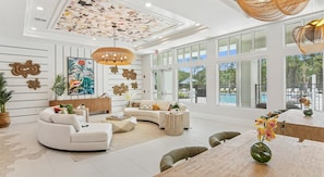 A spacious living room with a curved sofa, a coffee table, and a dining area with a wooden table and chairs. The room features large windows, a ceiling with a geometric pattern, and decorative wall art. - Landing | Chic 2BD, Pool, Clubhouse (Palm Bay)