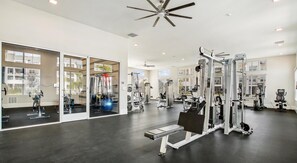 A spacious gym with various exercise equipment, large windows, and a ceiling fan. - Landing | Chic 2BD, Pool, Gym (Palm Bay)