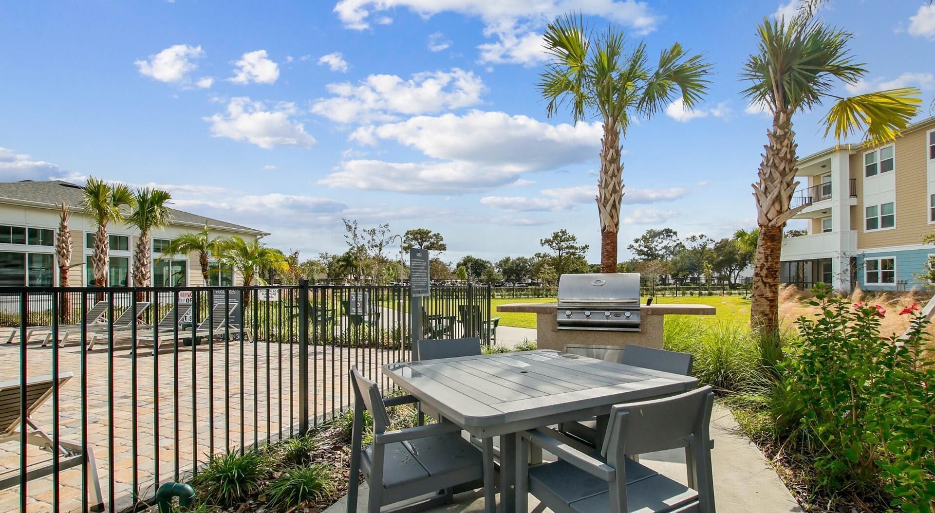 Un patio al aire libre con una parrilla, asientos y vista a un barrio residencial con palmeras y cielo despejado.