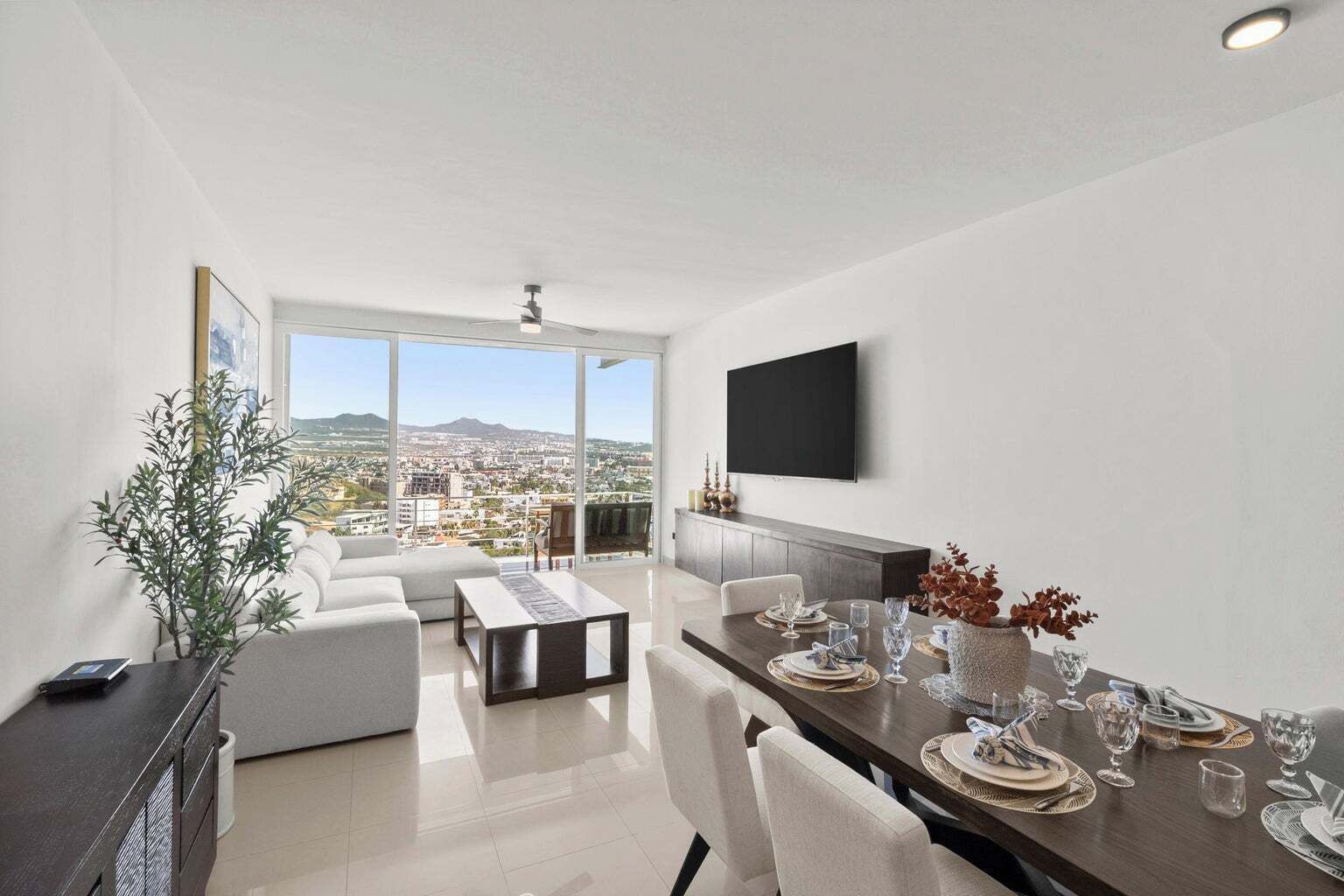 A modern living room with a dining area, large windows offering a city view, a flat-screen TV mounted on the wall, and a potted plant.