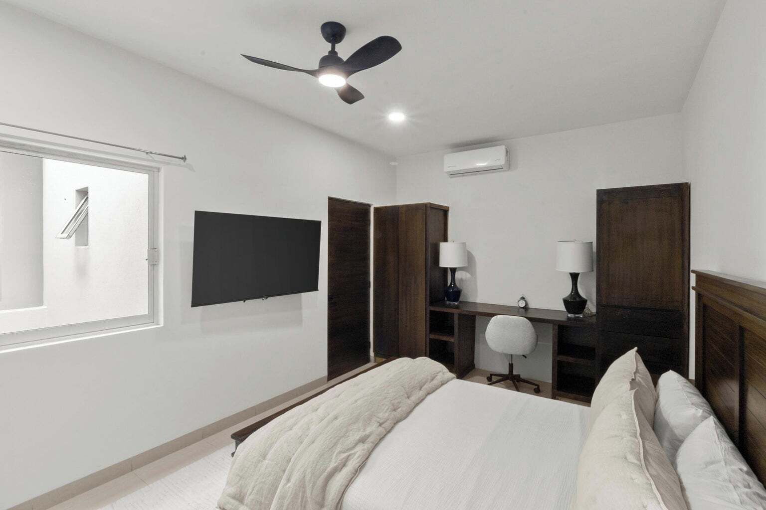 A bedroom with a bed, a ceiling fan, a TV, a desk with a chair, and a window.