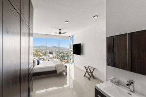 A modern bedroom with a bed, a TV, and a view of a cityscape. - NEW! Luxury Condo Pedregal Ocean and City Views, 15min Walk to downtown (Cabo San Lucas)