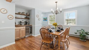 A dining area with a wooden table and chairs, a chandelier, and a window with a view of trees. - Peaceful Lakes Region Retreat  | Canoe + Fire pit (Meredith)