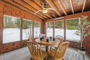 Eine Holzhütte mit rundem Tisch und Stühlen, großen Fenstern und einem Deckenventilator.