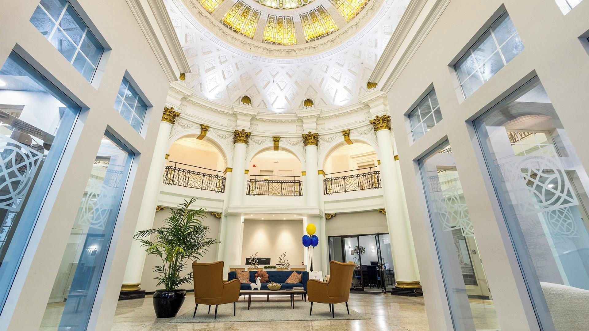 A spacious lobby with a high ceiling, large windows, and a central seating area with a plant and two chairs.